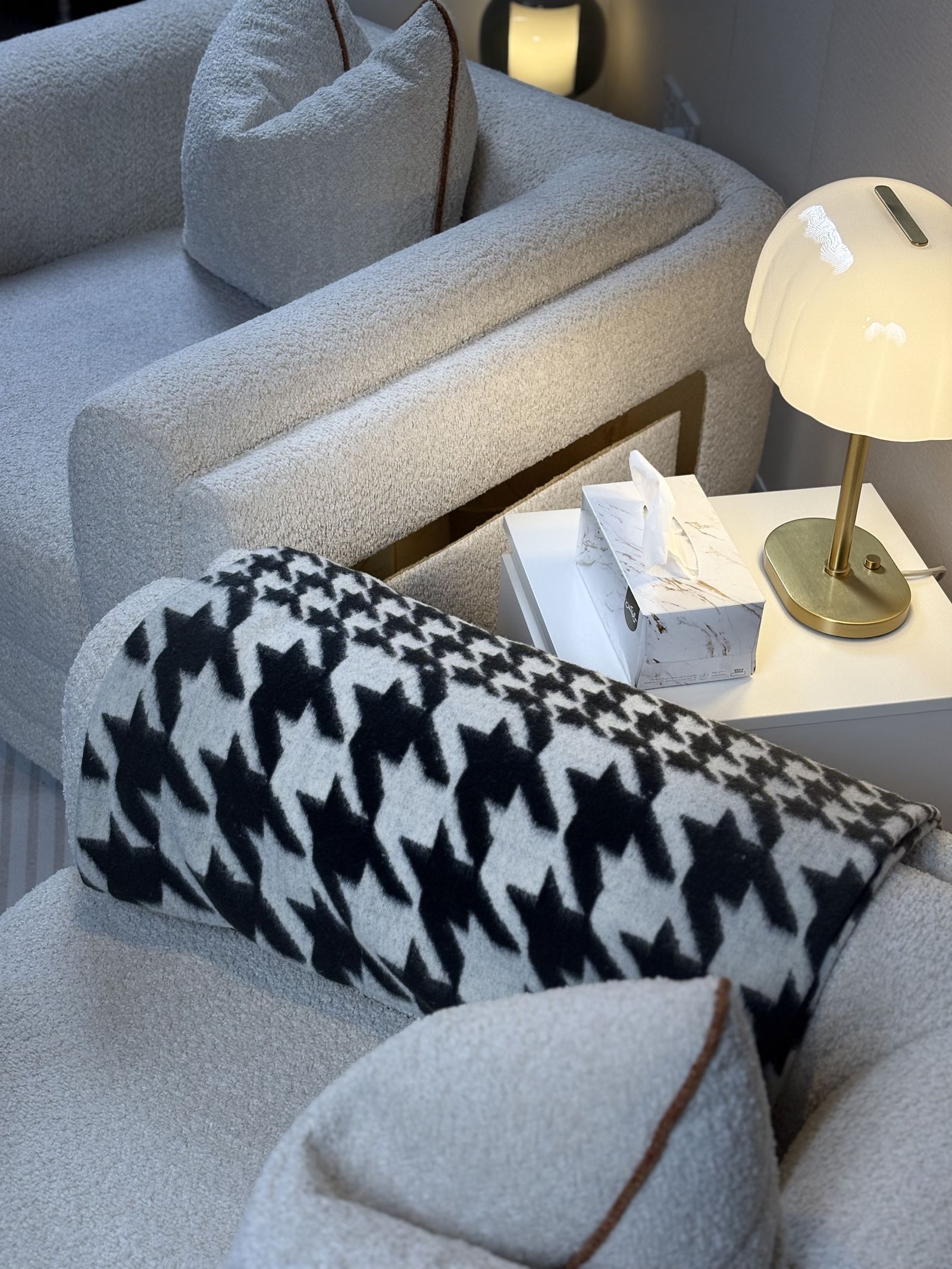 Interior detail: bouclé armchair with a houndstooth bolster, brass table lamp and marble tissue box, warmly lit
