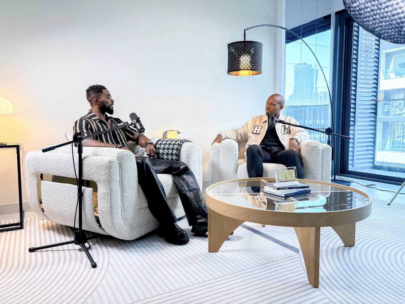Two guests seated in bouclé armchairs on a bright podcast set, boom microphones on stands, arc lamp overhead and floor-to-ceiling windows behind
