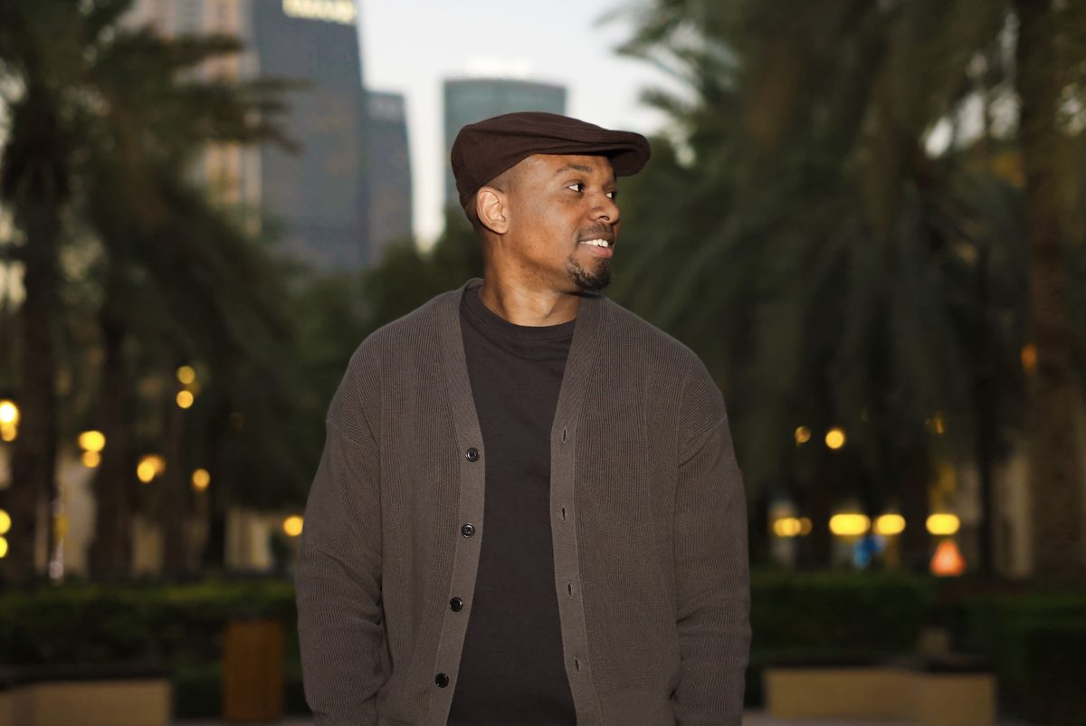 Portrait of a man in a flat cap and cardigan, palm trees and lit walkway behind him