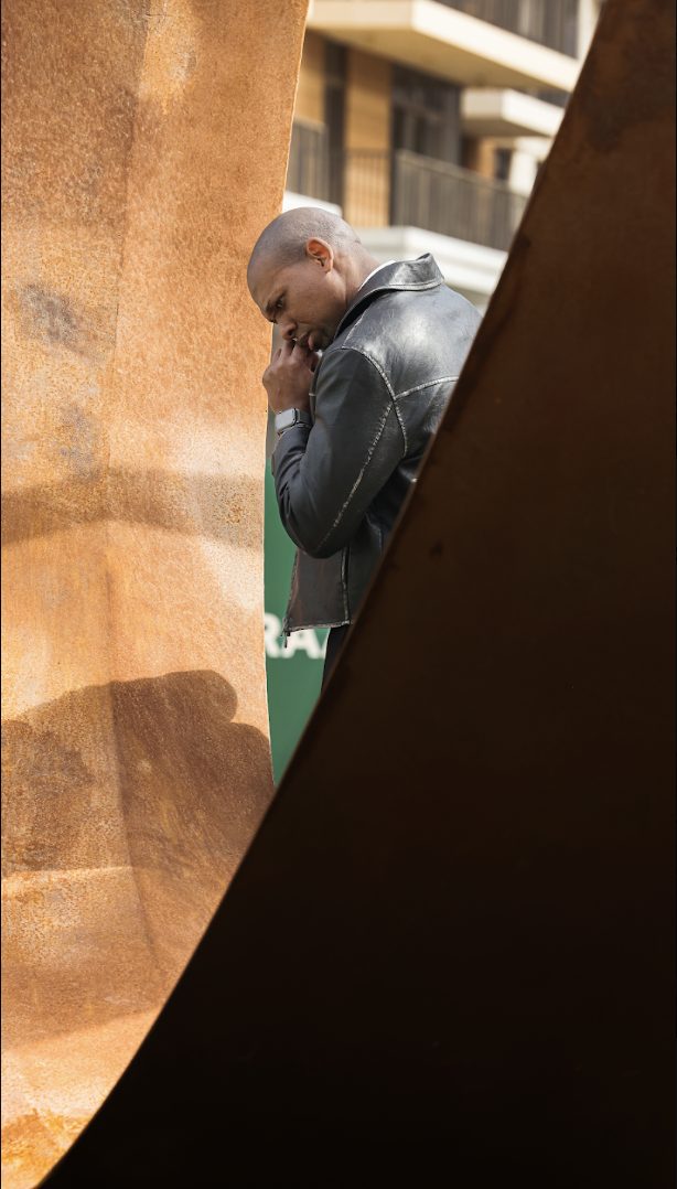 Editorial portrait of a man in a leather jacket framed between two curved weathered steel sculpture panels