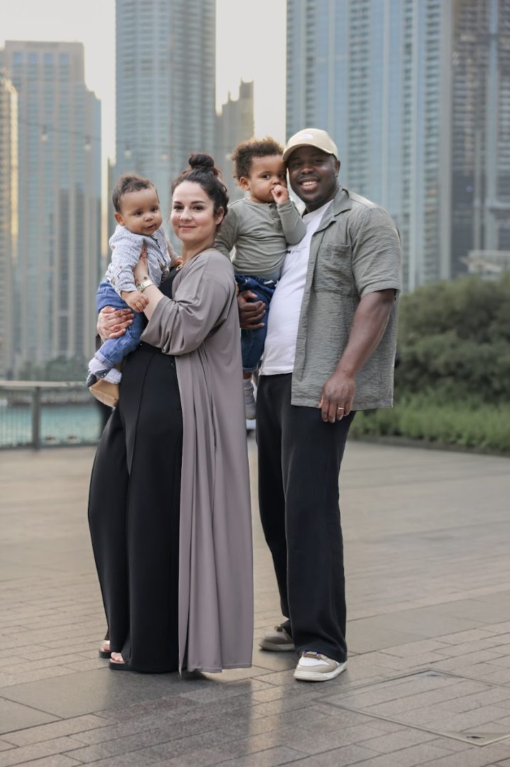 Family portrait: two parents holding toddlers, city towers and water behind them at dusk