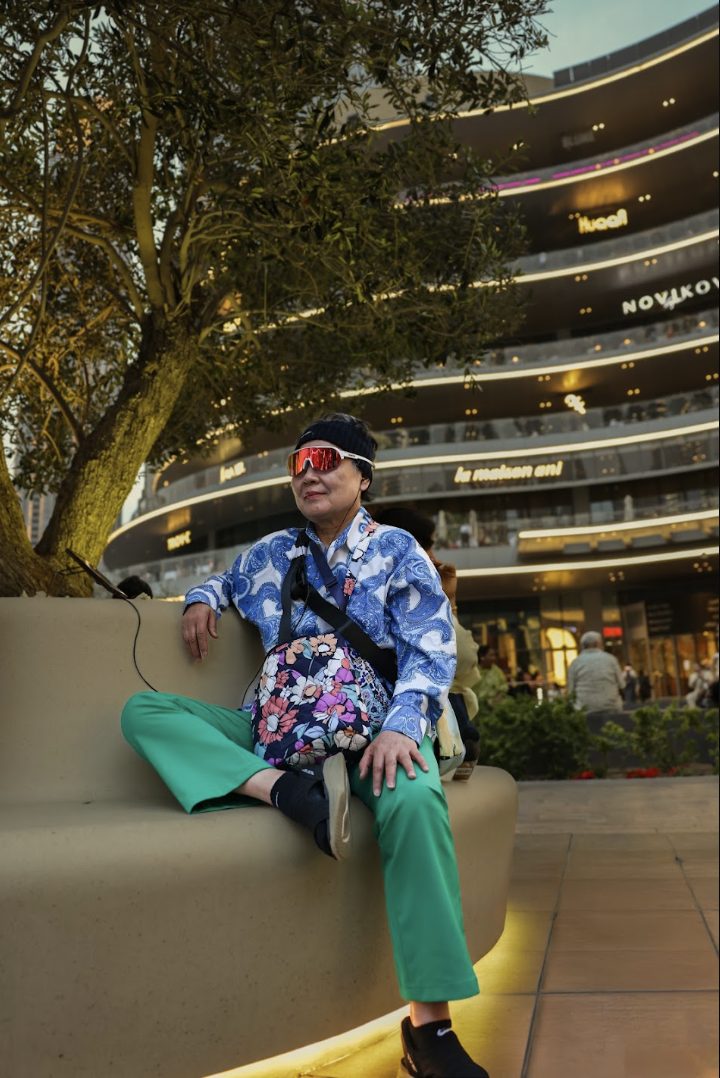 Street portrait: a woman in green trousers and a paisley shirt seated on a curved bench beneath an olive tree, illuminated building behind
